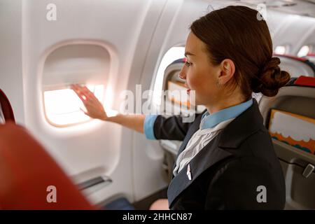 Donna assistente di volo chiusura finestra in aereo Foto Stock