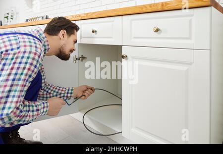 Giovane idraulico che ripara il lavandino e che usa un cavo di scarico per pulire un tubo ostruito Foto Stock