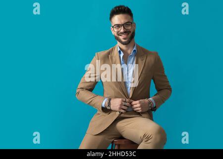 seduto felice uomo giovane con occhiali sorridenti e regolare vestito mentre seduto su sfondo blu in studio Foto Stock