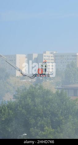 Vigili del fuoco su camion piattaforma che va sul tetto di casa a fuoco. Vista dei vigili del fuoco che cercano di estinguere il fuoco da edifici bruciati in fiamme e fumi. Uomini che arrestano lo smog e il fumo dalla casa. Foto Stock