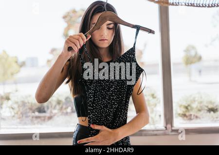 Giovane bruna che tocca e ispeziona l'abito punteggiato alla moda mentre ti trovi vicino alla finestra durante il giorno a casa Foto Stock