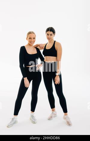 Due donne vestite in sport si legano l'una all'altra mentre si levano in piedi al coperto su sfondo bianco. Persone, esercizio e concetto di sport Foto Stock