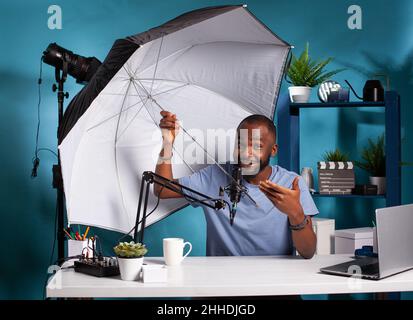 Fotografo professionista vlogger revisione flash riflettore ombrello presentare dimensioni e vantaggi in podcast online. Revisore di tecnologia che prova le apparecchiature dello studio di fotografia per vlog. Foto Stock