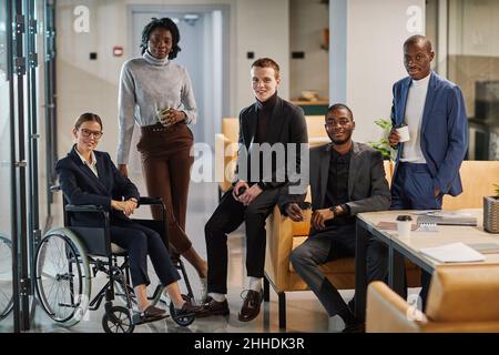 Ritratto completo di diversi team aziendali con una giovane donna in sedia a rotelle, tutti sorridenti alla macchina fotografica in ufficio Foto Stock