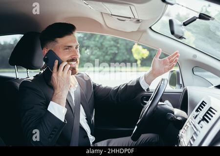 Giovane uomo con barba uomo d'affari che guida un'auto nel parcheggio sorridendo riuscito a parlare al telefono Foto Stock