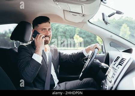 Giovane uomo con barba uomo d'affari che guida un'auto nel parcheggio sorridendo riuscito a parlare al telefono Foto Stock