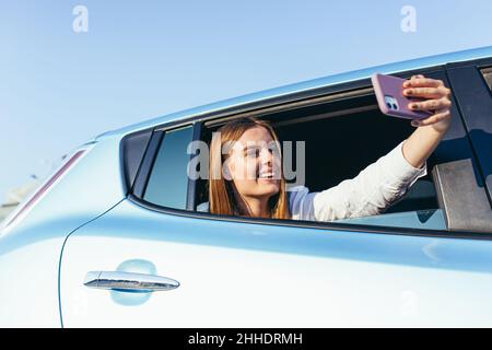 Giovane bella donna che parla di videochiamata seduto in auto attaccando la sua mano con il telefono fuori dalla finestra Foto Stock