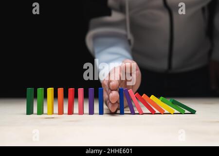 Una mano che raggiunge tra dominali allineate e colorate e quindi ferma una reazione a catena. Foto Stock