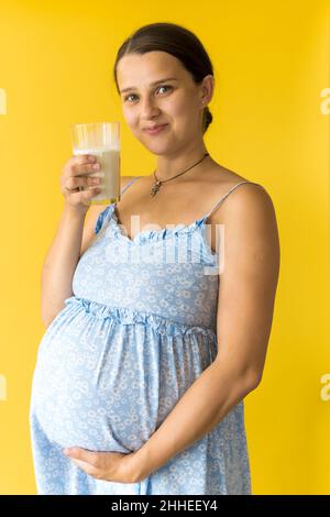 ritratto cute bella gravidanza giovane bella donna graziosa in abito blu floreale tiene un bicchiere da bere di latte strofina tummy su sfondo giallo. Maternità Foto Stock