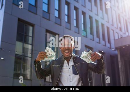 Un uomo d'affari di successo in abiti casual gioisce nel successo, African American guarda nella macchina fotografica sorride e tiene dollari in contanti Foto Stock