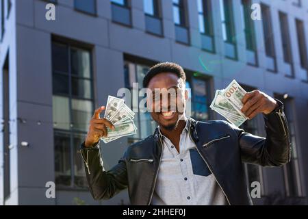 Un uomo d'affari di successo in abiti casual gioisce nel successo, African American guarda nella macchina fotografica sorride e tiene dollari in contanti Foto Stock