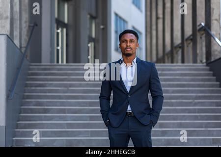 Ritratto giovane fiducioso successo afro-americano uomo d'affari guardando fotocamera serio uomo d'affari in piedi su sfondo urbano di ufficio moderno Foto Stock