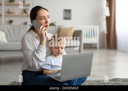 Madre affollata e stressata che parla sul cellulare mentre il figlio del bambino si siede sulle ginocchia della mamma e guarda il computer portatile Foto Stock