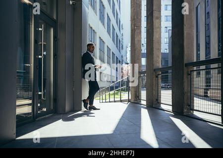 Uomo d'affari maschio vicino ufficio legge le notizie dal calcolatore del tablet, freelance afro-americano nel vestito di affari vicino ufficio fuori Foto Stock