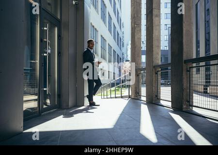 Uomo d'affari maschio vicino ufficio legge le notizie dal calcolatore del tablet, freelance afro-americano nel vestito di affari vicino ufficio fuori Foto Stock