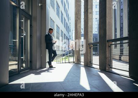 Uomo d'affari maschio vicino ufficio legge le notizie dal calcolatore del tablet, freelance afro-americano nel vestito di affari vicino ufficio fuori Foto Stock