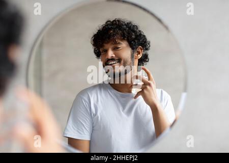 Felice uomo indiano guardando a specchio spruzzando su colonia in bagno interno, facendo mattina la routine di cura della pelle Foto Stock