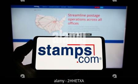 Persona che tiene il telefono cellulare con il logo della società di spedizione americana Stamps.com Inc. Sullo schermo di fronte alla pagina web aziendale. Mettere a fuoco sul display del telefono. Foto Stock