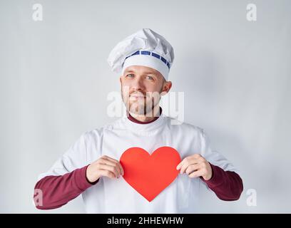 Chef attraente che tiene un cuore rosso davanti all'abbigliamento da lavoro. Chef o panettiere bearded in uniforme bianca con cuore di carta rossa come San Valentino, romanticismo o concetto di amore. Foto di alta qualità Foto Stock