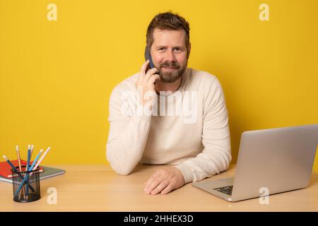 L'uomo allegro e giovane barbuto si siede e lavora alla scrivania con un computer portatile mentre parla su smartphone isolato su sfondo giallo. Foto Stock