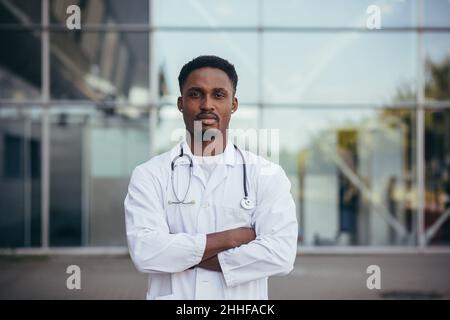 Ritratto di un giovane medico afroamericano serio, vicino alla clinica, in un abito medico bianco guardando la macchina fotografica Foto Stock