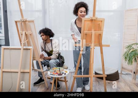 Donna afroamericana sorridente in piedi vicino a tela su cavalletto, ragazzo e vernici a casa Foto Stock