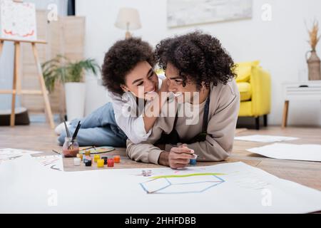 Positiva donna afroamericana abbracciante ragazzo con vernice vicino carta sul pavimento Foto Stock