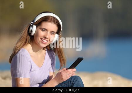 L'adolescente felice che indossa le cuffie che tengono il telefono cellulare vi guarda ascoltare la musica in natura Foto Stock