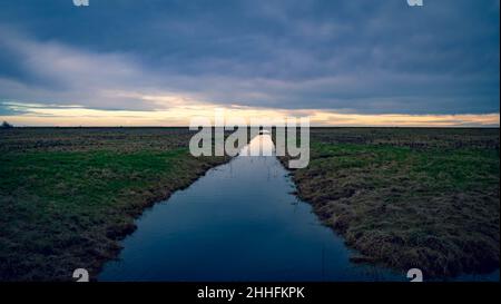 Freiston Shore, Boston, Lincolnshire. Le nuvole scure e l'orizzonte dorato luminoso riflesso in un uomo fermo hanno fatto il canale pieno d'acqua che si recava all'infinito. Foto Stock