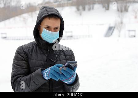 Protezione da coronavirus, uomo in maschera medica su una strada invernale della città con smartphone in mano. Concetto di sicurezza durante il covid-19 pandemia, tempo di neve Foto Stock