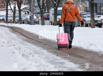 Una donna in viaggio cammina lungo un marciapiede lastricato invernale sgusciato di neve, portando una valigia su ruote. Spazio di copia. Foto Stock