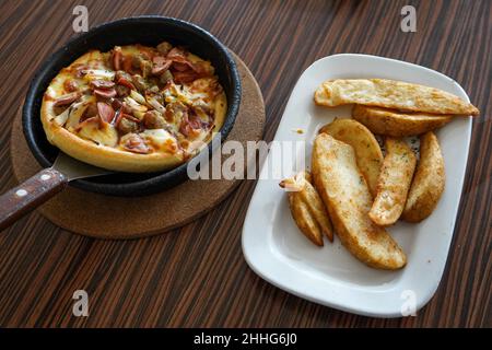 Barbeque beef pizza capanna servito su un piatto nero o ciotola con un marrone testurizzato tavolo di legno sfondo. Patate fritte condite servite su un piatto bianco. Foto Stock