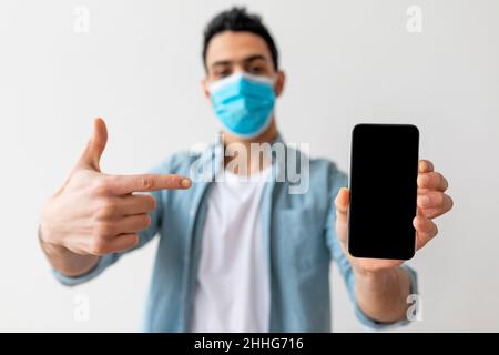 Giovane uomo arabo in maschera medica protettiva che punta lo smartphone con schermo vuoto, dimostrando mockup Foto Stock