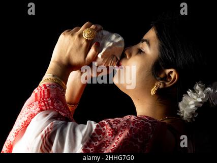 Una bella donna indiana in rosso saree soffiando conchiglia su sfondo nero Foto Stock