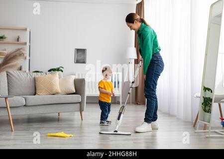 Madre giovane insegnando bambino bambino pulizia a casa, pulizia del pavimento in soggiorno interno, spazio libero Foto Stock