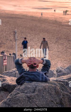 Un uomo seduto su rocce rilassandosi e guardando la luce tarda sera su Fistral a Newquay in Cornovaglia. Foto Stock