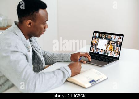 Formazione remota, brainstorming. Ragazzo astuto afro-americano, studente o freelance, si siede a un tavolo a casa in cucina, ha un webinar online, prende appunti, sullo schermo gruppo di persone multirazziali Foto Stock