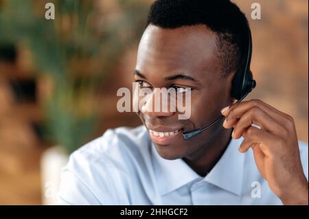 Primo piano di un simpatico uomo afro-americano con cuffie, esperto, consulente o operatore di hotline, rispondendo ai clienti chiamata, sorridente amichevole. Concetto di servizio di supporto, helpline, comunicazione remota Foto Stock