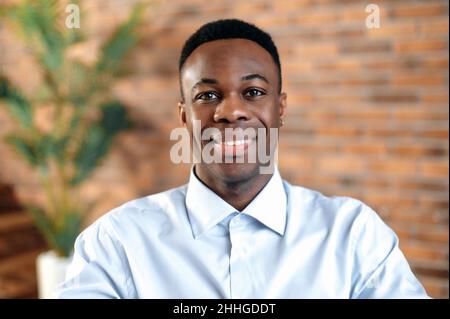 Primo piano di un bel uomo d'affari afroamericano sorridente, proprietario di piccola impresa, dirigente aziendale o direttore delle vendite, agente immobiliare di successo, in un ufficio moderno, guardando la macchina fotografica, sorridendo Foto Stock