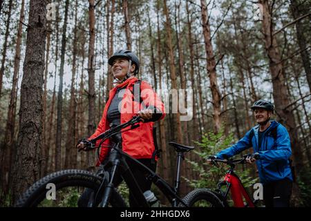 Vista ad angolo basso dei ciclisti senior che camminano e spingono le e-bike all'aperto nella foresta nel giorno d'autunno. Foto Stock