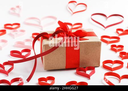 Scatola con regalo avvolta in carta kraft e legato con nastro rosso circondato da cuori di carta rossa su sfondo bianco Foto Stock