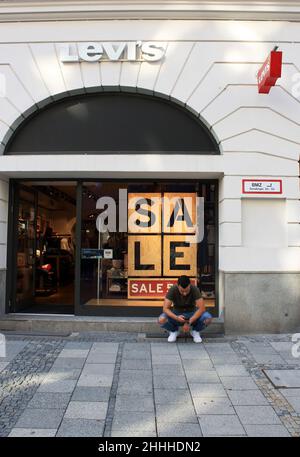 Monaco di Baviera, Germania - Luglio 7,2021:il negozio di abbigliamento 'Levis'brand, una vetrina con un'iscrizione 'Sale' e un uomo seduto vicino al negozio su croste, mi guarda Foto Stock