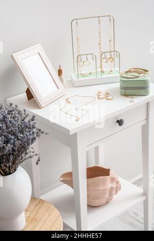 Tavolo con gioielli alla moda e vaso con fiori vicino alla parete luminosa Foto Stock