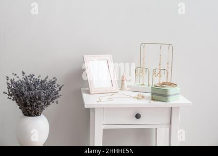 Tavolo con gioielli eleganti e vaso con fiori vicino alla parete luminosa Foto Stock