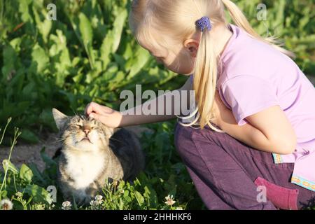 Adorabili poco ragazza caucasica accarezzare il suo gatto all'aperto alla luce del sole in estate. Foto Stock