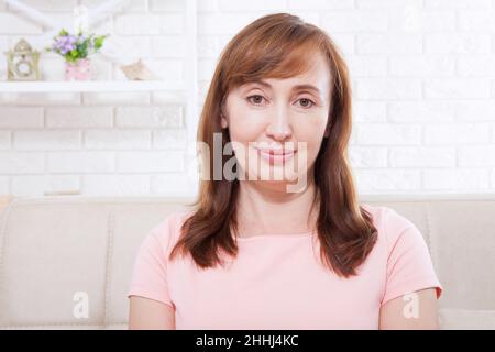 Sorridente bella bruna di mezza età donna seduta sul divano a casa guardando la macchina fotografica. Faccia con grinze. Menopausa. Copia lo spazio e riponi Foto Stock