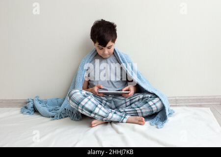 Un ragazzo di bruna, un adolescente in pigiama grigia, sul pavimento, sorride, usa un tablet digitale Foto Stock