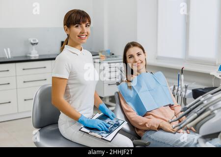 Dentista amichevole con tablet e immagini radiografiche che spiegano il trattamento al paziente durante l'esame dei denti. Moderna clinica dentale. Concetto di cura orale professionale, consultazione in studio di stomatologia. Foto Stock