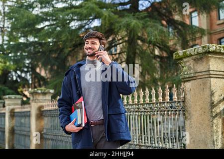 Allievo maschio allegro in outerwear in piedi con i notepads ed il piegatore ed avere conversazione telefonica sulla strada della città Foto Stock