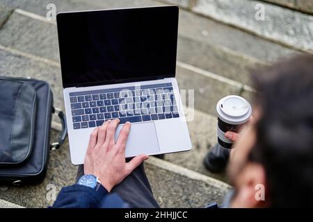 Vista posteriore ad angolo alto dello studente maschile seduto su scala di pietra e navigare netbook mentre si prepara per il compito universitario e avere caffè Foto Stock